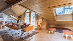 A cozy attic room with a wooden ceiling, a dining table with chairs, a sofa, and a window with a view of mountains.