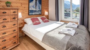 Hotellihuone, jossa on puinen seinä, valkoisilla lakanoiden ja punaisten tyynyjen peitetty sänky, vetolaatikoilla varustettu lipasto, vuoristoon avautuva ikkuna ja seinällä juliste.
