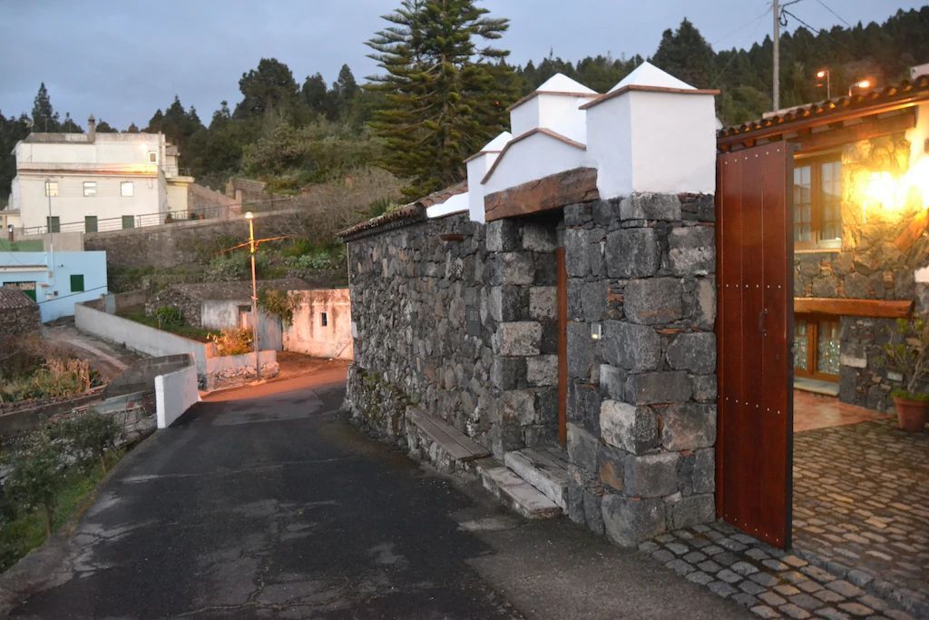 Un muro in pietra con una porta di legno, un vialetto lastricato e un edificio con facciata bianca sullo sfondo.