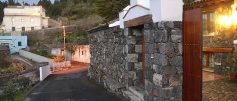 Un muro in pietra con una porta di legno, un vialetto lastricato e un edificio con facciata bianca sullo sfondo.
