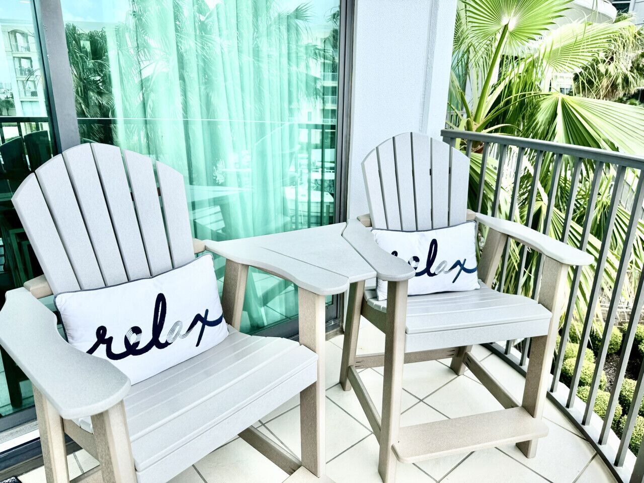 Two white Adirondack chairs with 'relax' pillows on a balcony with a glass railing and a view of palm trees.