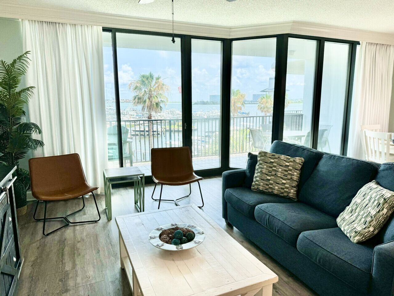 A modern living room with a sofa, two chairs, a coffee table, and large windows offering a view of a marina and palm trees.