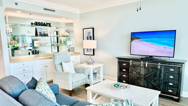Een woonkamer met een bank, fauteuils, een salontafel, een televisie en een keukenhoek met een aanrecht en kasten.