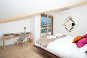 A bedroom with a bed, a desk, and a chair. There is a window with a view of houses and trees.