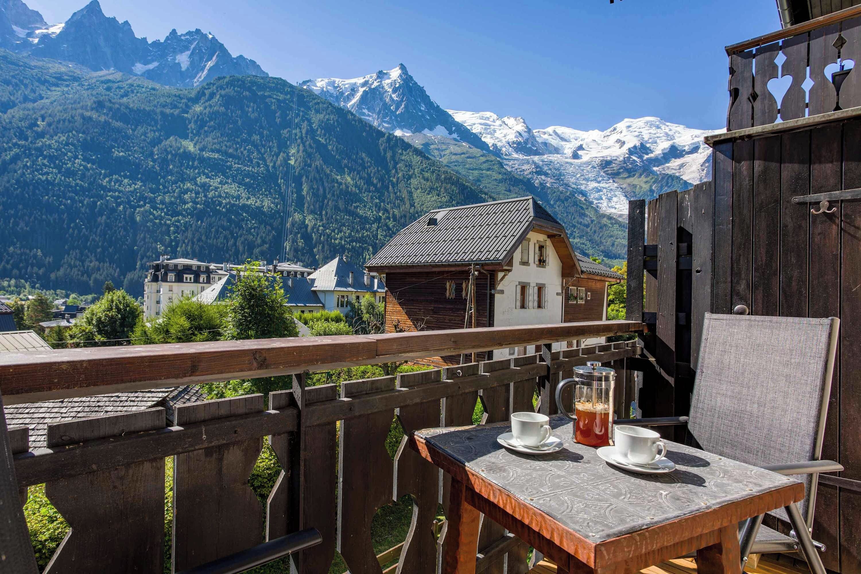 A balcony with a table and chairs, overlooking a mountainous landscape with snow-capped peaks and a village below.