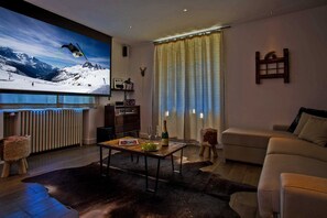 Een woonkamer met een tv met wintersportthema, een bank, een bijzettafeltje met een fles en glazen, en een bont tapijt.
