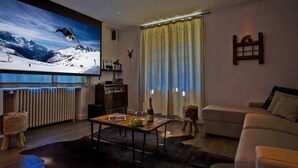 Una sala de estar con un televisor grande mostrando una escena de esquí, una mesa de centro de madera con una botella y vasos, un sofá y una alfombra de piel.