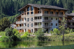 Un immeuble à plusieurs étages, doté de balcons et d’une façade en pierre, situé au bord d’un lac paisible, avec une forêt dense en arrière-plan.