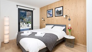 Een moderne slaapkamer met een bed, bedlampen, een rond bijzettafeltje en een raam met uitzicht op bergen.