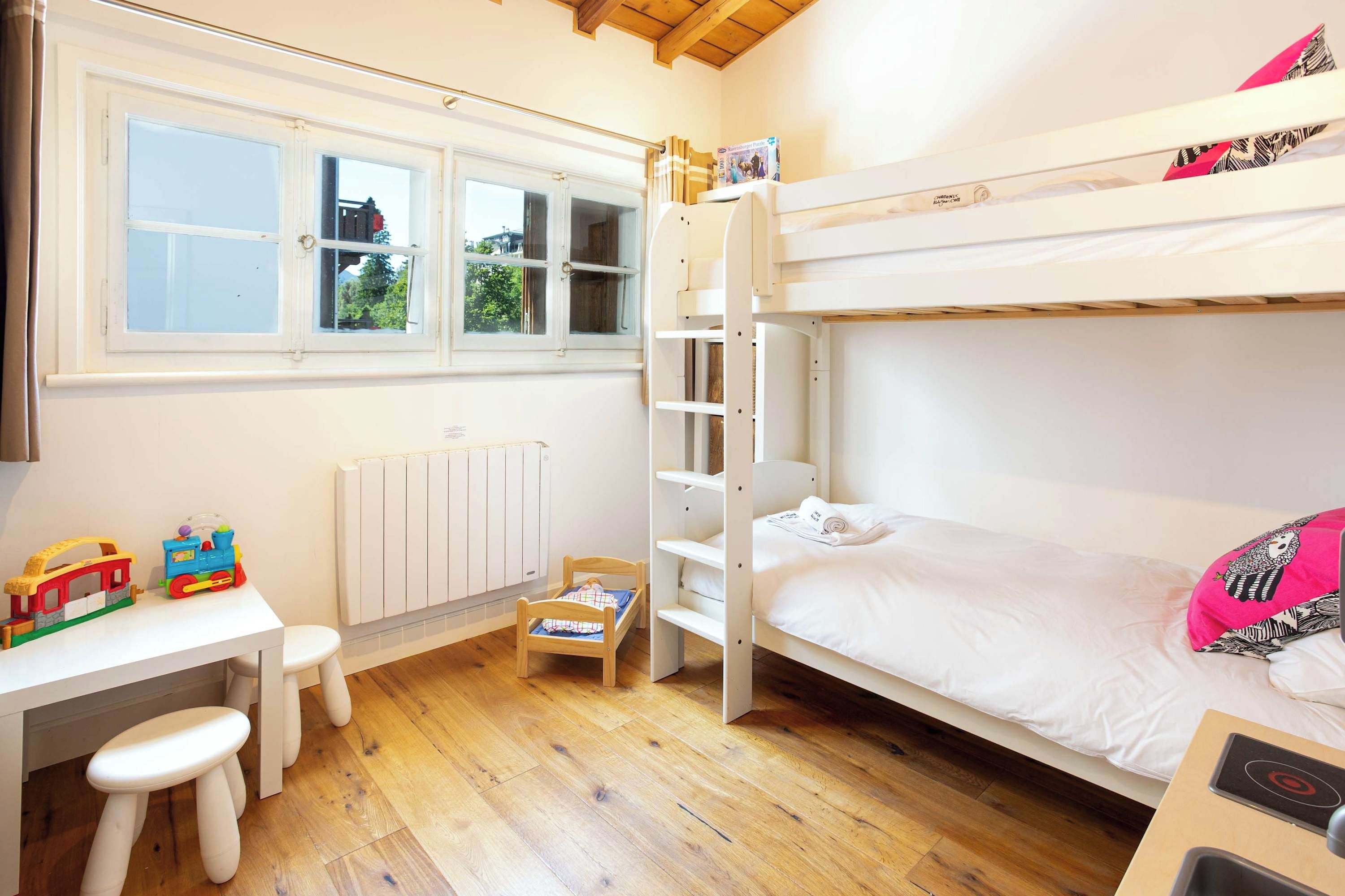 Quarto de criança com beliches, escrivaninha, radiador e janela com vista para o verde.