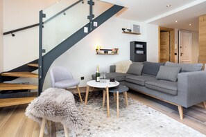 A modern living room with a grey sofa, a small round table, and a fluffy rug. A staircase with glass railings leads to an upper level.