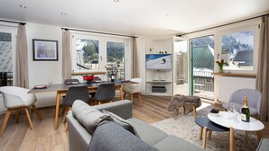 Un soggiorno moderno con divano, tavolo da pranzo e sedie. C'è una TV su un mobile, una finestra con vista sulle montagne e un balcone con ringhiera.