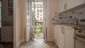 Una cocina estrecha con armarios blancos, un salpicadero de azulejos y vista a un edificio que dice 'HOTEL RICH' a través de una puerta abierta.