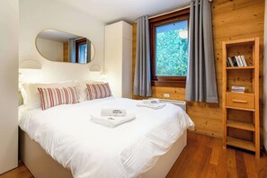 A hotel room with a bed, a bedside table, a mirror, a window with curtains, and a wooden bookshelf.