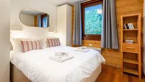 Ein ordentlich bezogenes Bett mit weißen Leinen und gestreiften Kissen, ein Holzregal mit Büchern, ein großer Spiegel und ein Fenster mit Vorhängen.
