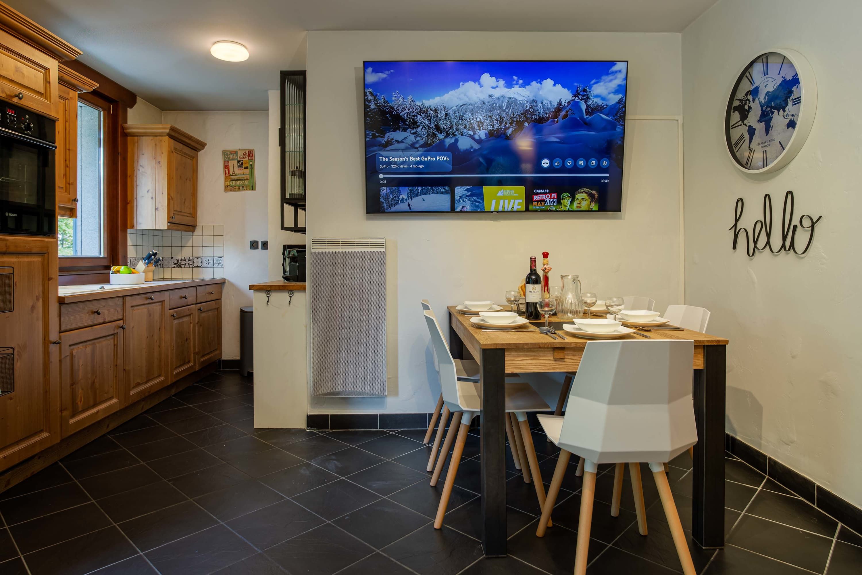 A modern kitchen with wooden cabinets, a dining table set for four, and a mounted television displaying a program.