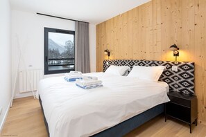 A modern bedroom with a large bed, wooden panel walls, a view of mountains through a window, and a bedside table with lamps. - La Cordee 612 Apt, Lakefront, Chamonix All Year (Les Praz)