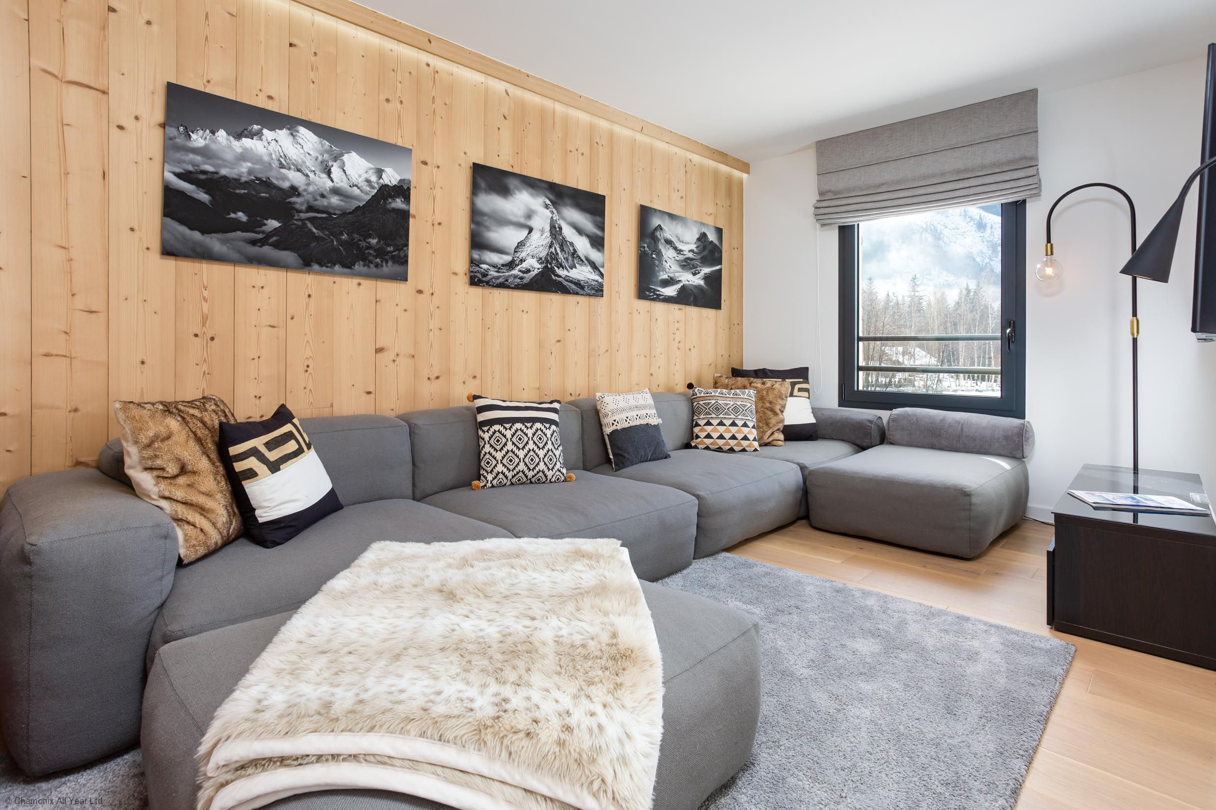 Un salon moderne avec un canapé d’angle, un plaid en fausse fourrure et trois photographies encadrées accrochées au mur.