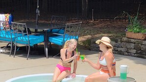 A swimming pool with two people sitting on the edge, a table with chairs, and a covered area with chairs. - Cozy N. Raleigh Apt. with Pool and Hot Tub. Pets welcome BBQ Grill, TV & movies (Raleigh)