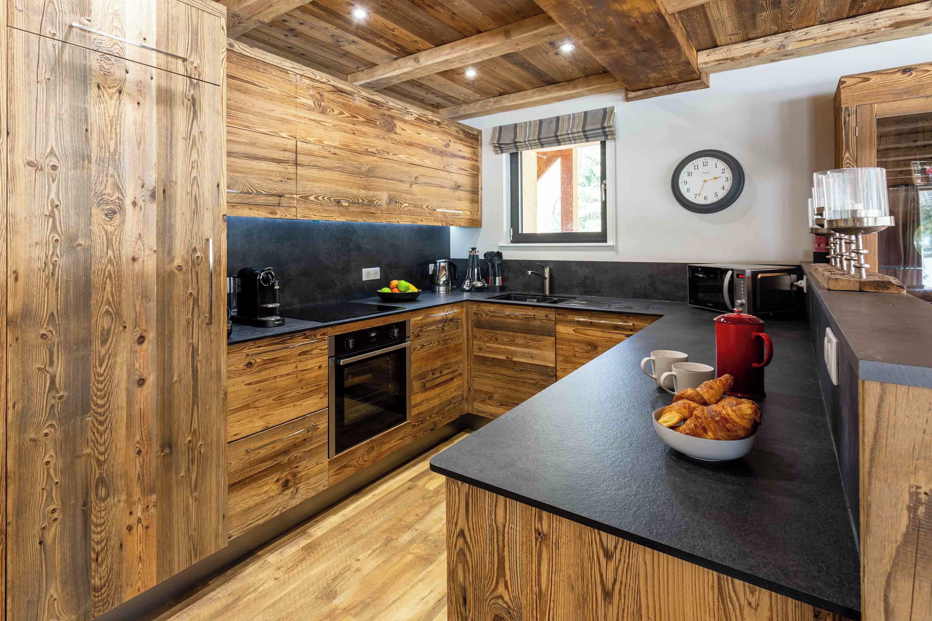 Une cuisine avec des meubles de rangement en bois, un plan de travail noir, une machine à café, une bouilloire et une horloge au mur.