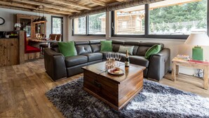 Un salon chaleureux avec un plafond en bois, de grandes fenêtres, un canapé d’angle en cuir, une table basse en bois et un petit coin cuisine.