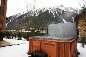 A hot tub with a wooden exterior, set against a snowy landscape with mountains in the background.