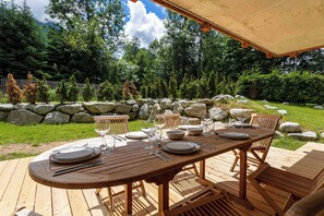 An outdoor wooden deck with a table set for dining, surrounded by a stone wall and greenery.