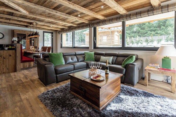 A modern living room with a wooden ceiling, a leather sectional sofa, a wooden coffee table, and a view of the outdoors through large windows.