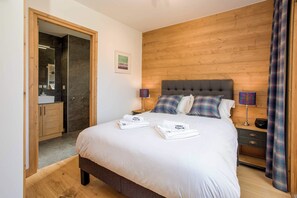 A hotel room with a large bed, wooden headboard, two bedside tables with lamps, a wooden wardrobe, and a bathroom visible through an open door.