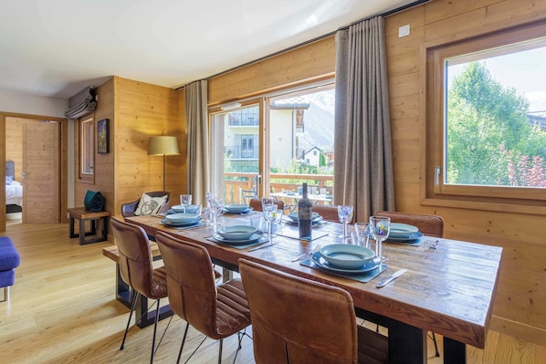 A dining area with a wooden table set for four, chairs, and a view of a mountain landscape through large windows.