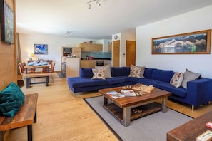 A modern living room with a blue sofa, a wooden coffee table, and a dining area with a wooden table and chairs. - Coin des Drus appt, Golfing, - Chamonix All Year (Les Praz)