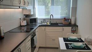 Ett modernt kök med vita skåp, en svart bänkskiva, ett matbord med duk, tallrikar och bestick, och ett fönster med utsikt över grönområden utanför.