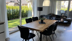 Un comedor moderno con una mesa de madera y sillas negras, ventanas amplias con cortinas, una lámpara de pie negra y un sofá negro al fondo.