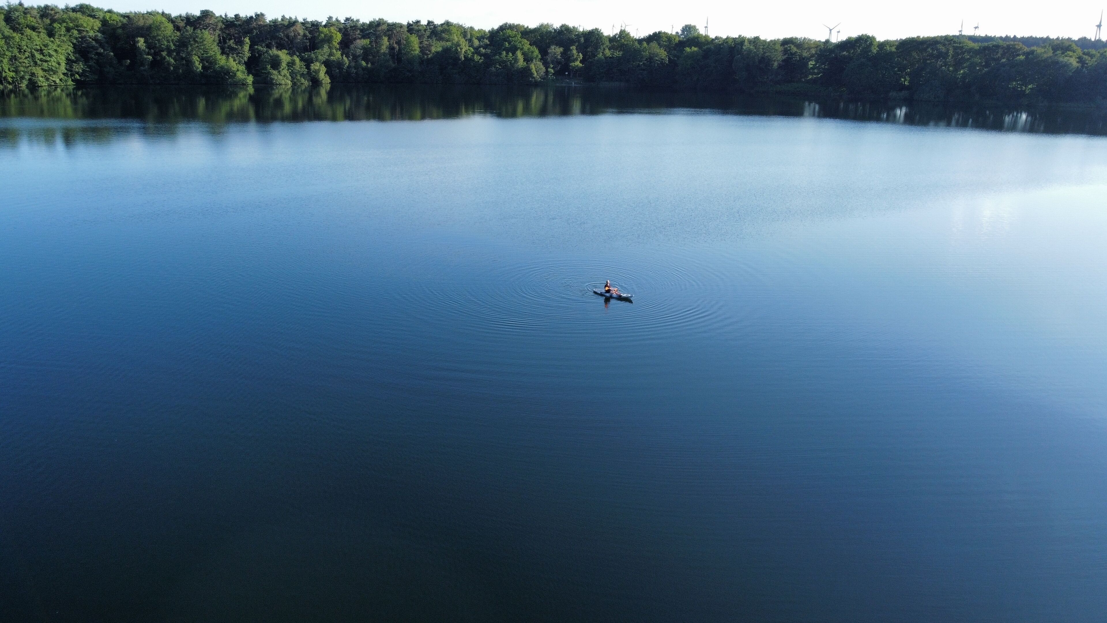 Um lago calmo com uma pessoa remando num caiaque, rodeada por árvores densas e turbinas eólicas ao longe.