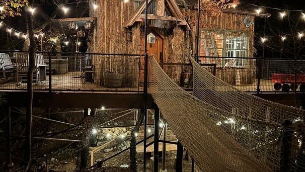 Een rustiek houten gebouw met een schuin dak, een touwbrug en lichtslingers.