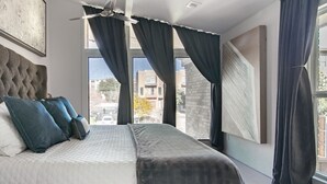 A modern bedroom with a large bed, dark curtains, a ceiling fan, and a city view. - High-Spec Opulent Townhouse in Oak Lawn (Dallas)