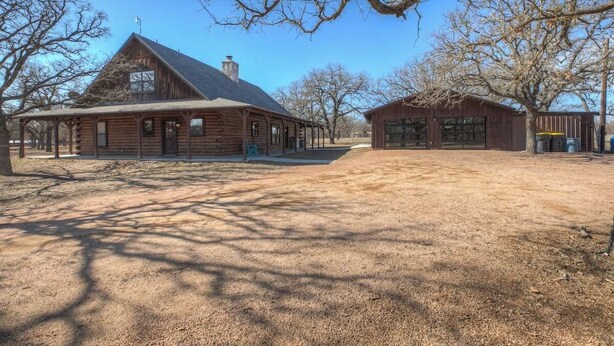 A rustic log cabin with a large porch, a separate building with a garage, and a fenced area with trees.