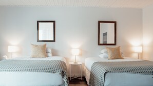 A hotel room with two beds, each with a patterned bedspread, a small bedside table with a lamp, and a framed mirror on the wall. - Kent Motel Niagara on the Lake (Niagara-on-the-Lake)