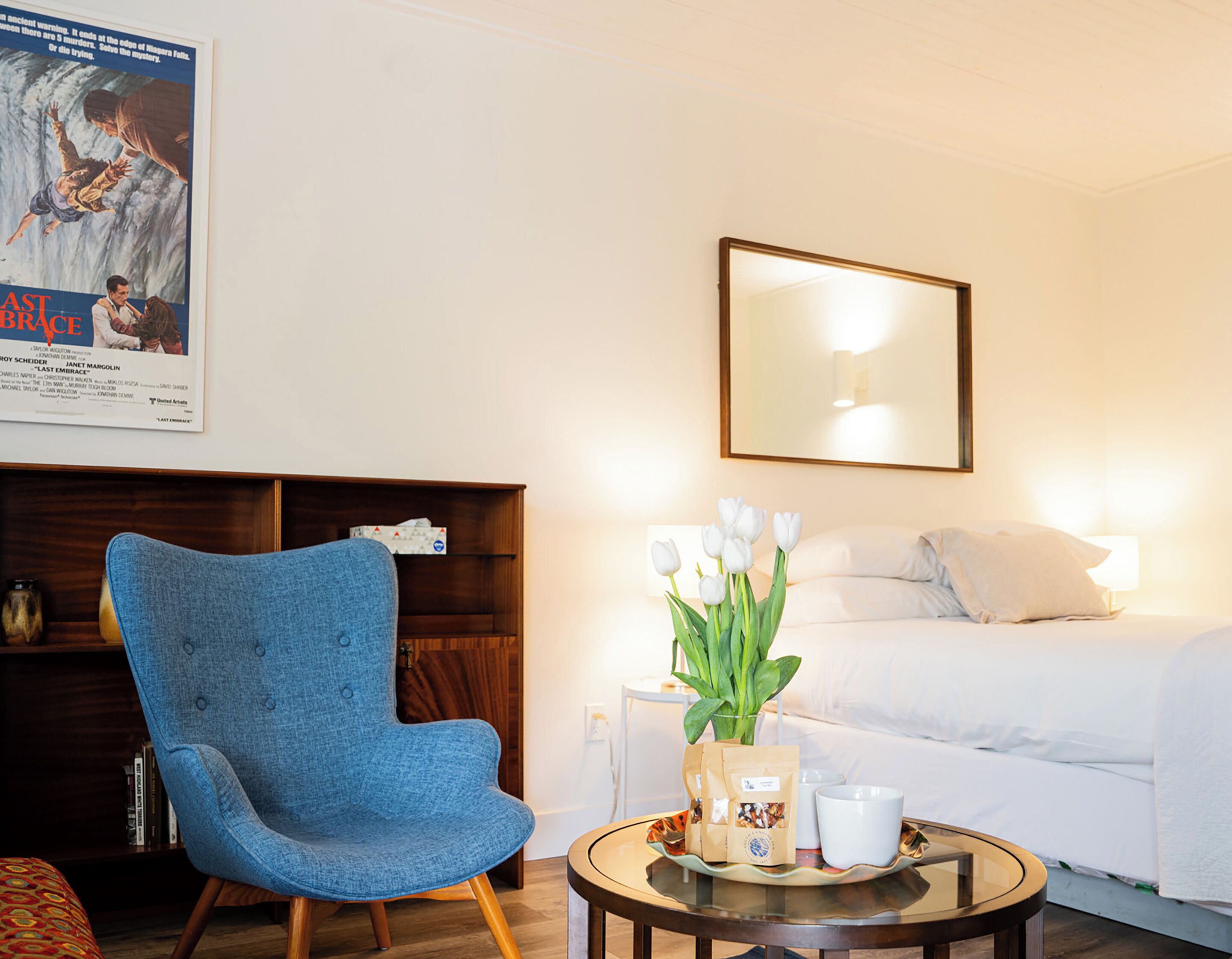 A hotel room with a bed, a blue armchair, a small round table with a tray, a vase with white flowers, and a poster on the wall.