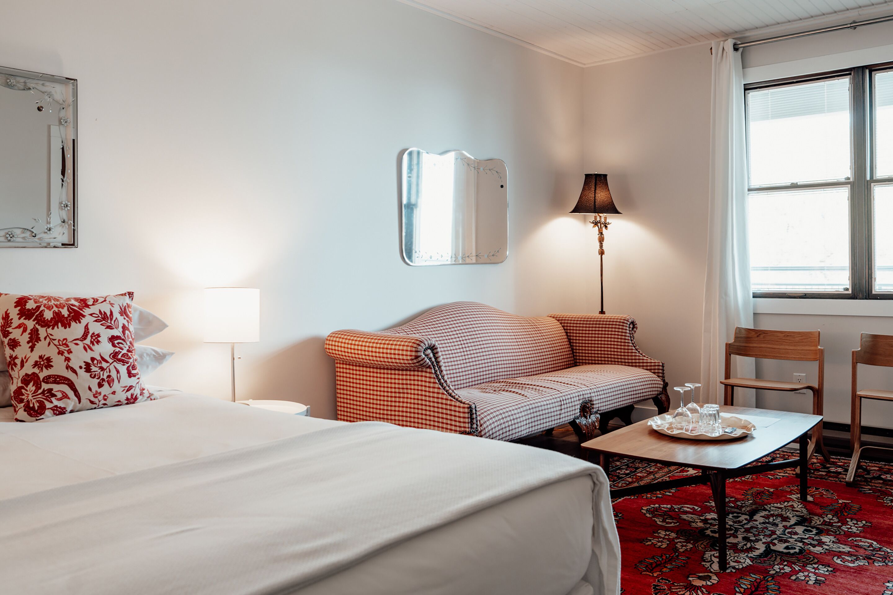 A hotel room with a bed, a sofa, a small table with a tray, a lamp, and a window with curtains.