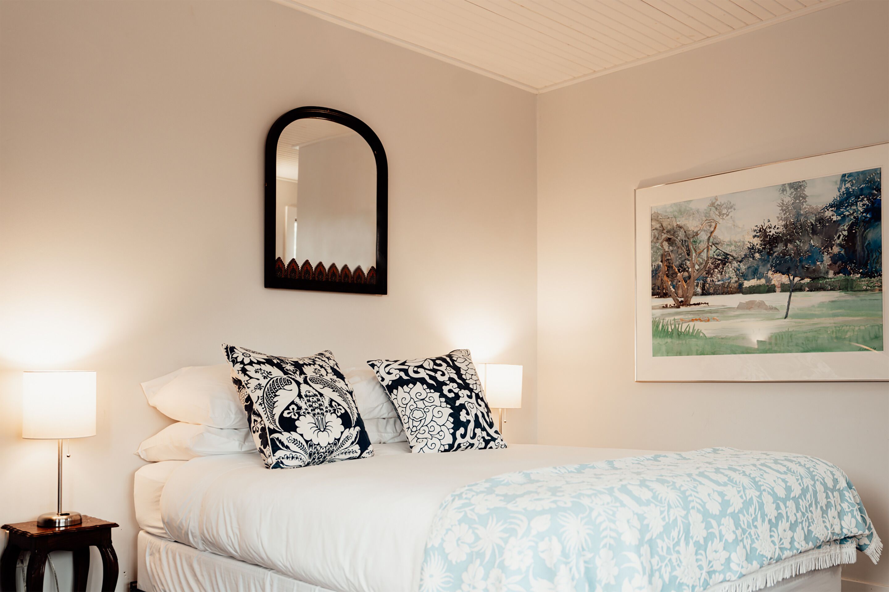 Una cama bien hecha con almohadas estampadas, una lámpara de noche, un cuadro enmarcado y un espejo en la pared.