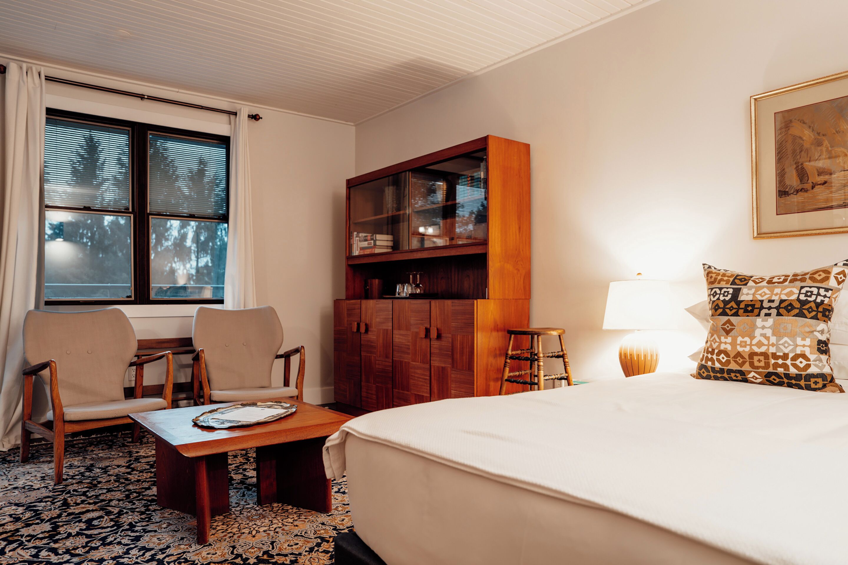 A hotel room with a bed, two chairs, a wooden cabinet, a table, and a window with curtains.