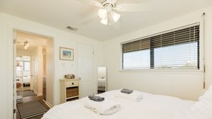 A bedroom with a bed, a ceiling fan, a window with blinds, a door, and a small cabinet. - Pineapple by the Sea - Outdoor Bar, Walk to Beach! (Jamaica Beach)
