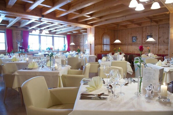 Een eetgedeelte met meerdere tafels gedekt voor een formeel evenement, met witte tafellakens, kaarsen en bloemstukken als decoratie.