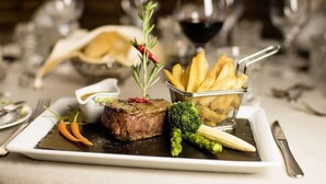 Ein angerichtetes Essen mit Steak, Pommes und Gemüse, im Hintergrund Weingläser.