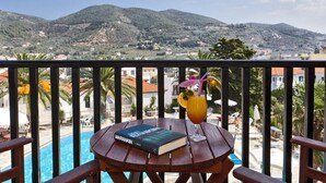 Una mesa de madera con un libro y un cóctel en un balcón con vista a una piscina y montañas.