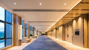 A long, well-lit corridor with large windows on one side, a patterned carpet, and a series of doors on the other.