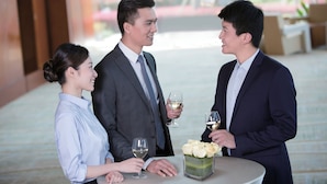 Three people at a round table with a vase of flowers, in a well-lit indoor setting with a view of outdoor greenery.