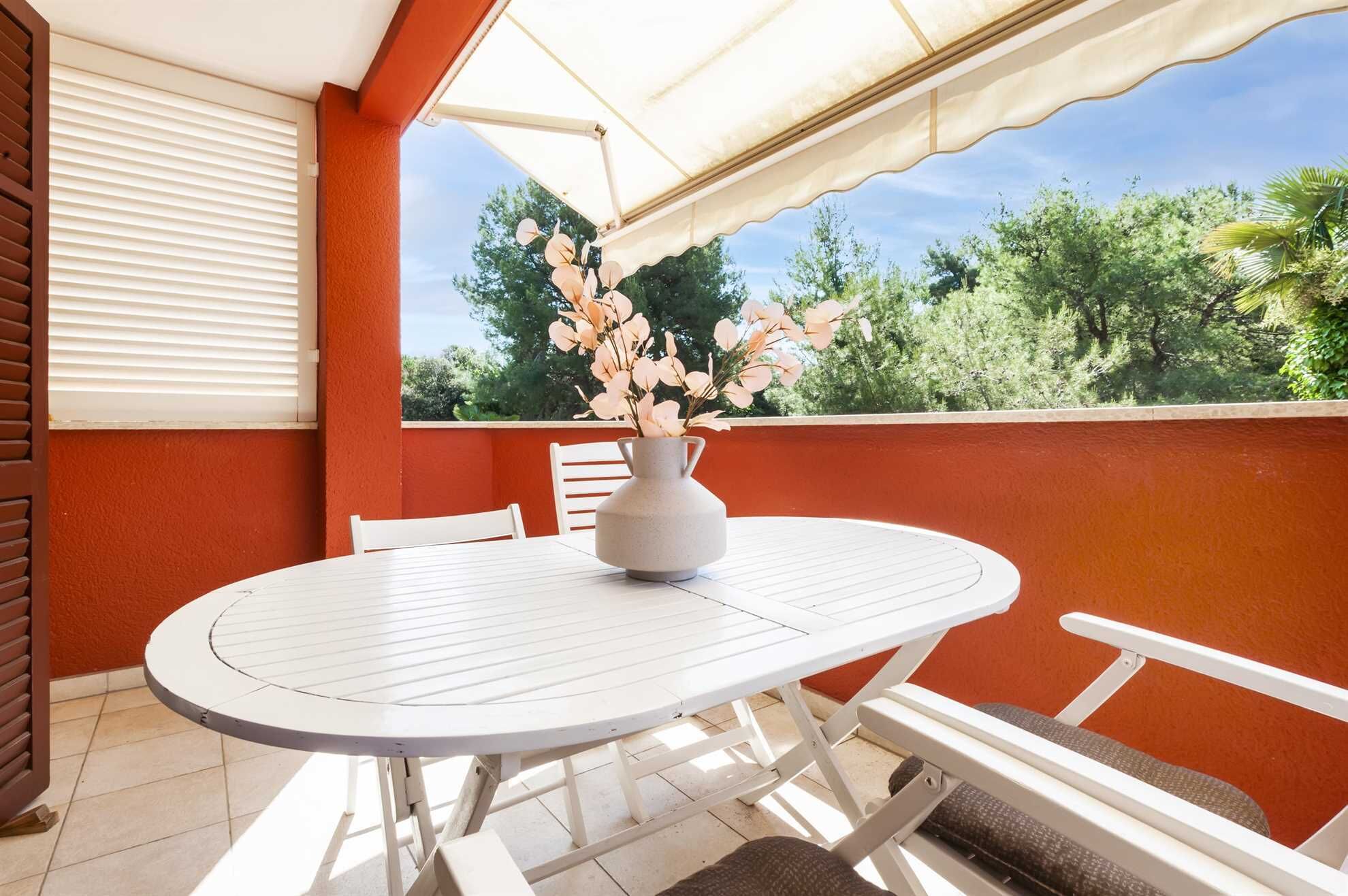 A round white table with a vase of flowers, white chairs, and a view of trees through a window with blinds.
