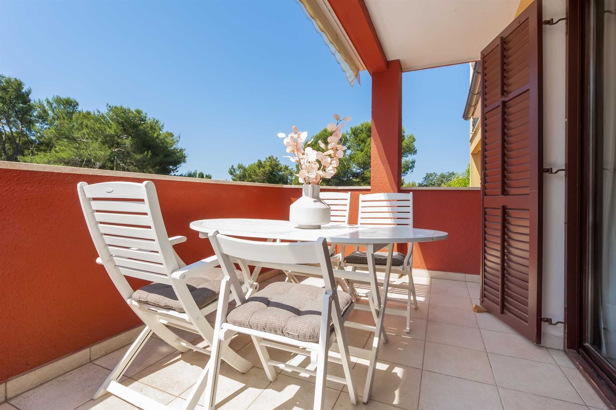 A balcony with a white table and chairs, a vase with flowers, and a view of trees.